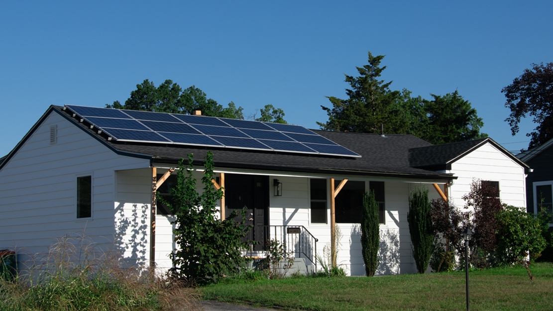 A single-story ranch-style home with white siding and a dark gray roof. On the roof, solar panels have been installed. The sky behind the home is bright blue.